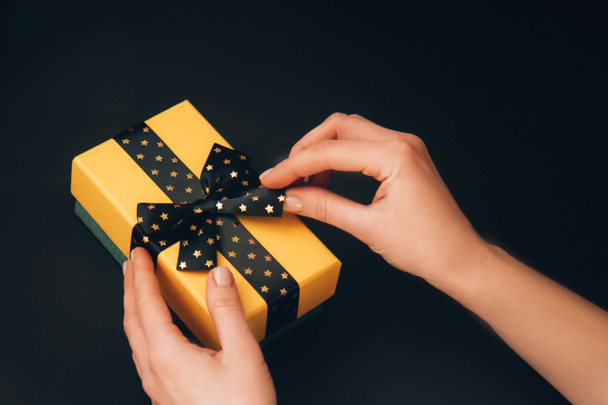 Boîte cadeau jaune avec arc noir avec des étoiles dorées dans les mains de la femme sur un fond sombre - Photo, image