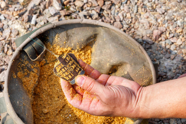 In the hand of a fisherman a feeder with fish food on the background of a canvas fishing bucket in blur and sunlight. Copy space, close-up. - Foto, Imagen