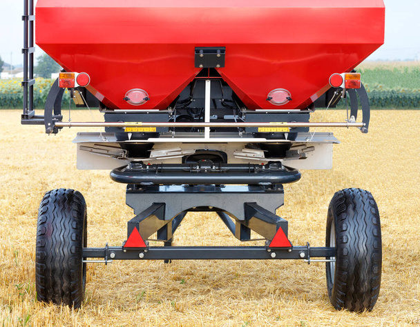 A red fertilizer spreader in the form of a drawbar stands against the background of a field of yellow straw harvested. Copy space. - Photo, image