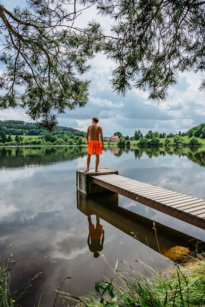 Rückansicht eines Mannes, der mit Freude von einem Holzsteg ins Wasser springt, die Natur spiegelt sich im See wider. Sommerspaß im Teich.Reiseurlaub, Jugendferienkonzept.Glücklicher aktiver Lebensstil, Unterhaltung. - Foto, Bild
