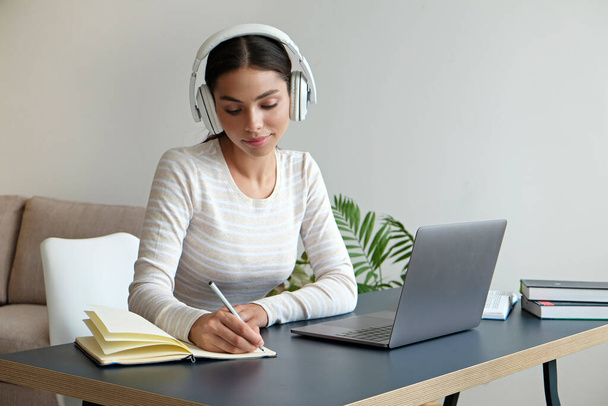 Online education concept. Young brunette woman wearing eyeglasses and headphones taking part in a group video call. Portrait of female college student studying at home. Close up, copy space background - Φωτογραφία, εικόνα