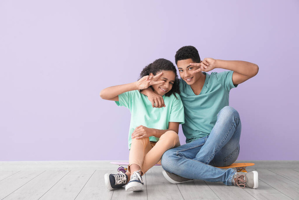 African-American brother and sister with skateboards showing victory gesture while sitting near color wall - Photo, Image