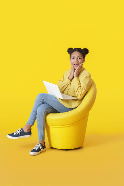 Überraschtes afroamerikanisches Mädchen mit Laptop sitzt im Sessel auf farbigem Hintergrund - Foto, Bild