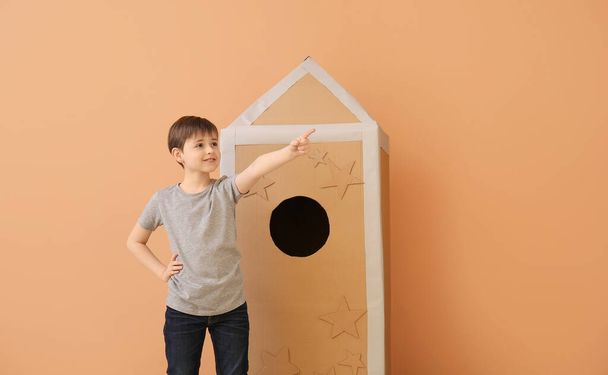 Schattig jongetje met kartonnen raket wijzend naar iets op kleur achtergrond - Foto, afbeelding