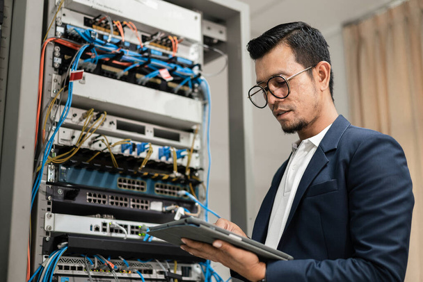 Stock photo of a young network technician holding tablet working to connecting network cables in server cabinet in network server room. IT engineer working in network server room - Photo, Image