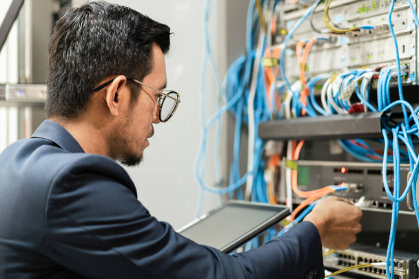 Складское фото молодого сетевого техника, держащего планшет, работающего над подключением сетевых кабелей в серверном шкафу в серверной сети. IT-инженер, работающий в серверной сети - Фото, изображение