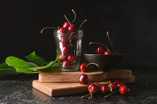 Bowl and jar with sweet cherry on dark background - Photo, Image