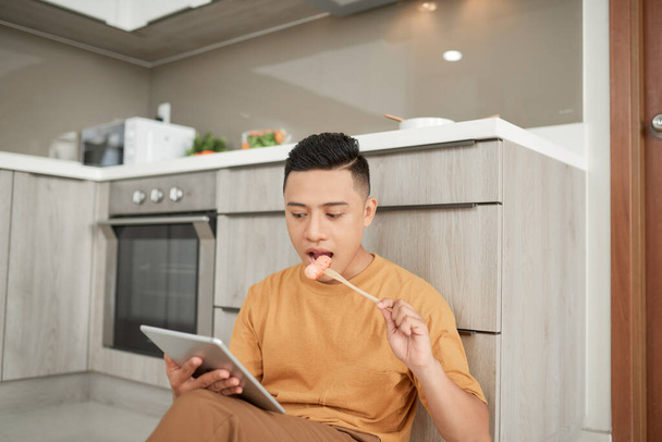 Νεαρός άνθρωπος χρησιμοποιώντας ψηφιακή ταμπλέτα κάθεται στο πάτωμα τρώει λουκάνικο στην κουζίνα - Φωτογραφία, εικόνα