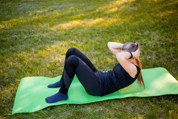 Mujer joven y delgada haciendo ejercicio abdominal mientras está acostada en una alfombra en el parque. - Foto, Imagen