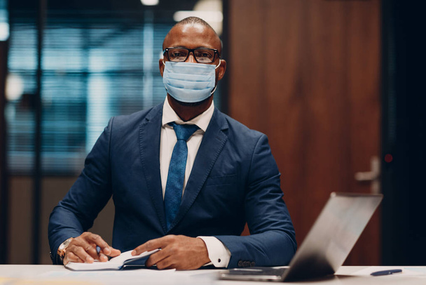 Portrait smiling african american businessman in medical face mask sit at table for meeting in office with notebook with pen and laptop. - Foto, Imagen