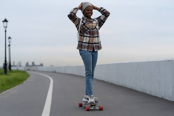 Fröhliches afrikanisches Mädchen, das mit aufgeregtem Lächeln Longboard reitet, genießt Freizeitaktivitäten im Freien - Foto, Bild