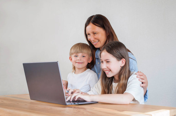 Mãe feliz com dois filhos estão sentados em casa na mesa com um laptop. Aprendizagem à distância de crianças. Mãe ajuda as crianças a aprender remotamente - Foto, Imagem