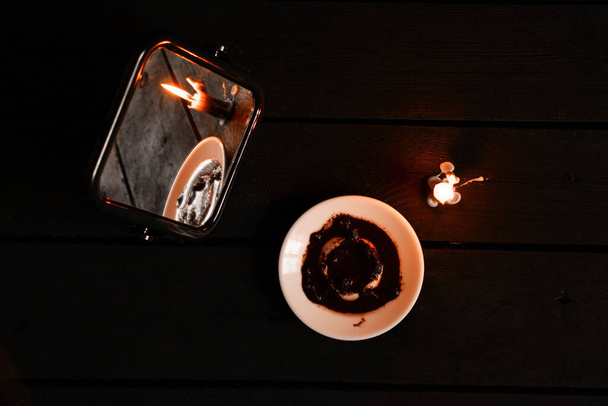 ένα αναμμένο κερί μεταφέρθηκε από καθρέφτες και υπάρχουν κάρτες που λένε την τύχη και τους λόγους του καφέ σε ένα κοντινό πιάτο, τύχη λέει στα χαρτιά από το φως των κεριών σε ένα μαύρο τραπέζι - Φωτογραφία, εικόνα