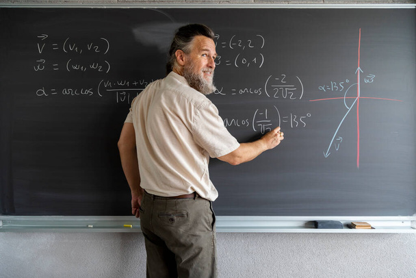 Homme caucasien mature professeur de lycée écrit des formules mathématiques sur le tableau noir avec de la craie. Espace de copie. Concept d'éducation. - Photo, image