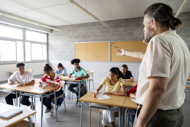 Mature adult caucasian man teacher stands in front of multicultural high school students giving an explanation. Copy space. Education concept. - Photo, Image