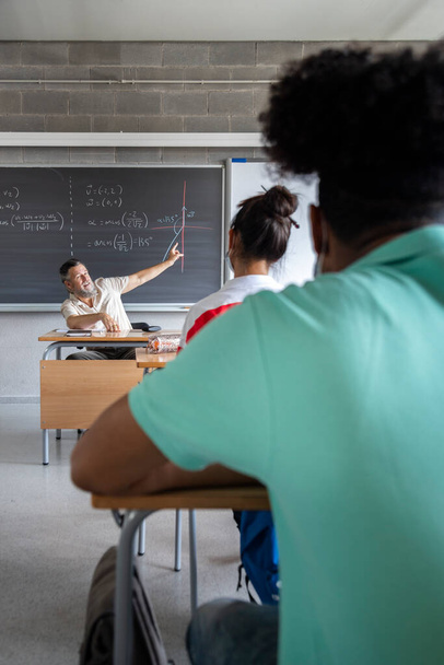 Caucasian mature man teacher with beard pointing blackboard during math class. Vertical image. Education concept. - Fotó, kép