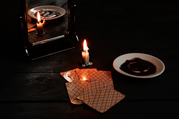 una vela encendida fue llevada por espejos y hay tarjetas de adivinación y café molido en un plato cercano, adivinación de la fortuna en tarjetas a la luz de las velas en una mesa negra - Foto, Imagen