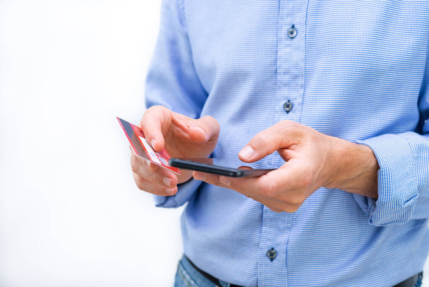 Man with phone and credit card in hands and blue shirt on white background. Copy space. - Φωτογραφία, εικόνα
