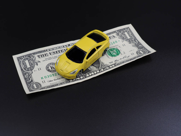Buying or renting a car. Car sharing. Yellow toy car on dollar bills on a black gradient background. Collapse of prices, discount on the car. - Photo, Image