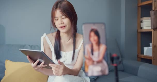 Happy young Asian girl blogger in front of phone camera use tablet enjoy question answer with follower in living room at home. Blogger activity lifestyle, Social distance coronavirus pandemic concept. - Metraje, vídeo