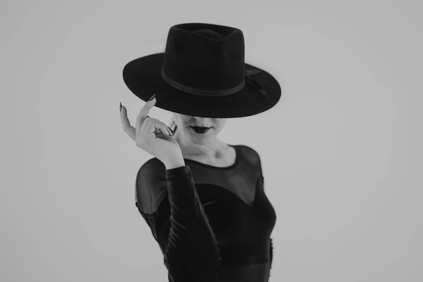 Чарівна жінка позує з чорним капелюхом на білому тлі. Femme fatale костюм, стильний одяг. Чорно-білий. Сексуальна модель танцюриста
. - Фото, зображення