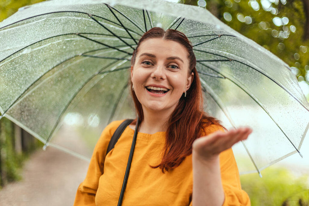Una bella donna sta sotto un ombrello trasparente e stende la mano, catturando gocce d'acqua. Bandiera orizzontale, spazio di copia - Foto, immagini
