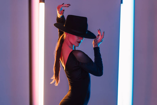 Alluring woman in hat between flashing colored neon lights, studio background. Femme fatale, stylish outfit of ballet dancer. - Foto, imagen