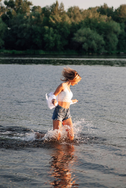 Photo of a running blonde in profile. A young woman in a white top and shirt and denim shorts runs freedom on the water against the background of a beautiful landscape at sunset - Fotó, kép
