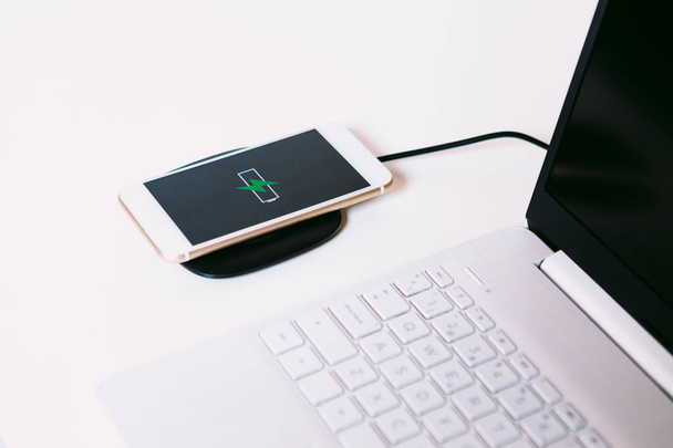 Білий мобільний смартфон з логотипом батареї з зеленою блискавкою на екрані, зарядка на базі зарядного пристрою без кабелю поруч з ноутбуком на білому робочому столі
 - Фото, зображення