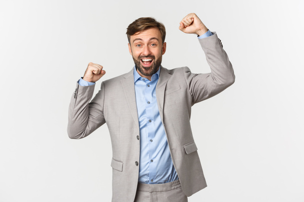 Portrait of successful smiling businessman with beard, wearing grey suit, cheering and celebrating victory, saying yes or hooray, triumphing and winning, standing over white background - Photo, Image