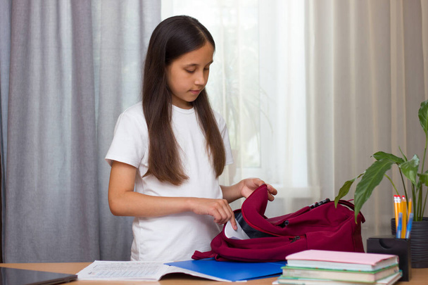 A girl with dark long hair in a white T-shirt stands at the table at home and puts her notebooks into a school backpack. Back to school concept - 写真・画像