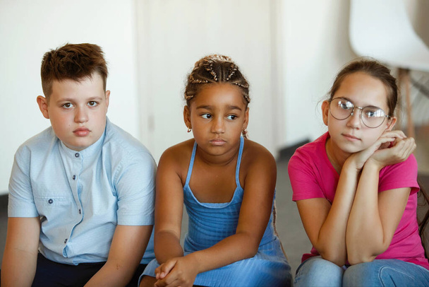 Τρεις λυπημένοι, σοβαροί έφηβοι κάθονται. Καυκάσιος, Αφροαμερικανός, αγόρι και κορίτσι με γυαλιά. - Φωτογραφία, εικόνα
