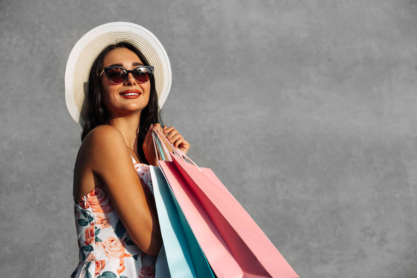 Joyeux beau jeune femme en chapeau d'été, porte robe et lunettes de soleil tient des sacs isolés sur fond gris, concept de shopping - Photo, image