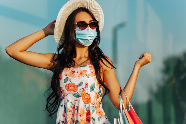 Портрет щасливої молодої жінки в медичній захисній масці від вірусів, з сумками біля торгового центру в місті, споживання, покупки, коронавірус, карантин
 - Фото, зображення