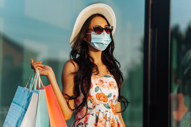 Portrait of a happy young woman in a medical protective mask from viruses, with bags near a shopping center in the city, consumerism, shopping, coronavirus, quarantine - Photo, image