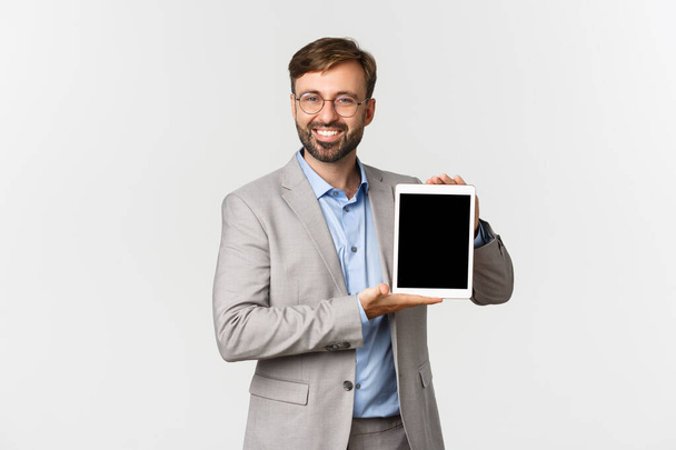 Изображение успешного и уверенного в себе предпринимателя в сером костюме и очках, демонстрирующего что-то на цифровом планшетном экране и улыбающегося, стоящего на белом фоне - Фото, изображение