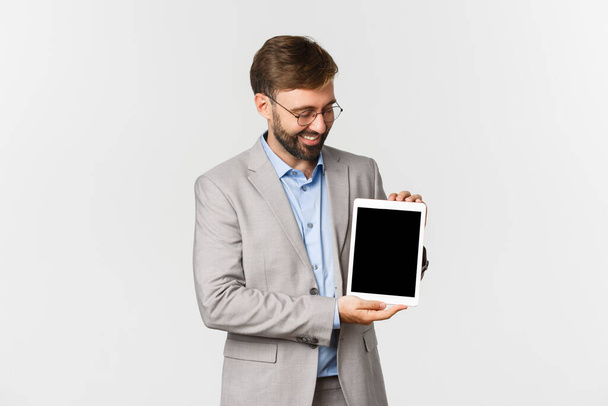 Portrét šťastného a spokojeného podnikatele v šedém obleku a brýlích, hledící a ukazující digitální tablet, hrdě se usmívající, stojící nad bílým pozadím - Fotografie, Obrázek