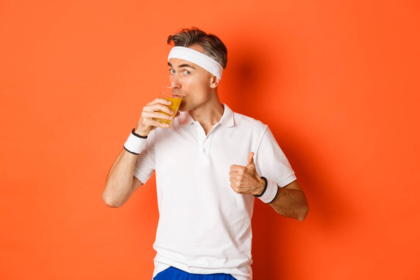 ワークアウト、ジム、ライフスタイルの概念。ハンサムな中年のフィットネス男の肖像,親指アップと飲酒ジュースを示します,オレンジの背景の上に立って - 写真・画像