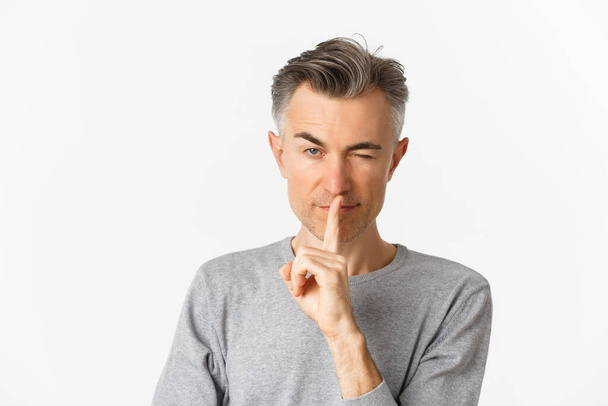 Großaufnahme eines frechen gutaussehenden Mannes mittleren Alters, der zwinkert und Schweigezeichen setzt, um Geheimhaltung oder Schweigen bittet, vor weißem Hintergrund steht - Foto, Bild