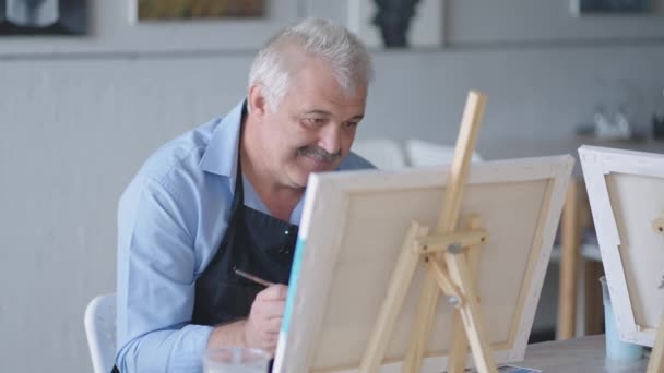 Eine Lehrerin zeigt einem pensionierten Mann bei Kursen für Senioren, wie man mit Farben und Pinsel ein Bild zeichnet. Ein älterer Mann zeichnet einer Rentnergruppe ein Bild - Filmmaterial, Video