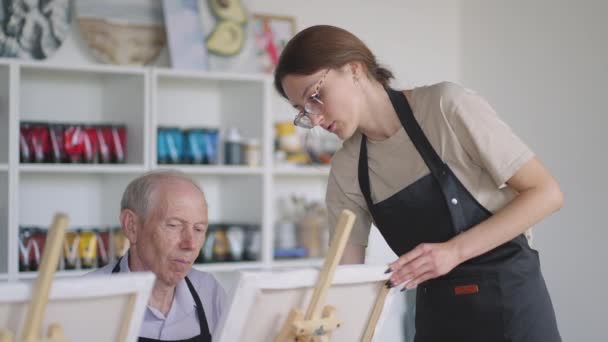 Una maestra le muestra a un hombre jubilado cómo dibujar un cuadro con pinturas y un pincel en los cursos para ancianos. Un hombre mayor dibuja un cuadro a un grupo de pensionistas - Imágenes, Vídeo