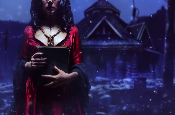 Witch or sorceress with runic makeup and wooden animal skull amulet holding a magic book standing in dark mysterious forest with hut on the background. Halloween concept, black magic. - Photo, Image