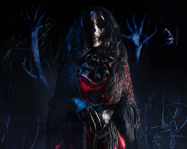Geisterfrau oder Hexe mit schrecklichem Make-up im gotischen Gewand, schwarzem Schleier und Jabot auf gruseligem Hintergrund mit Hexenhänden im Wald. Halloween-Konzept. - Foto, Bild