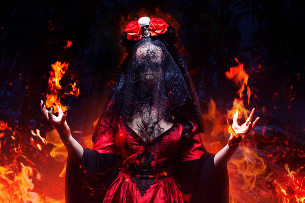 Heks in gotische jurk, zwarte bruidssluier en kroon met schedel en rozen die vuurspreuk uitgieten in het nachtbos. Halloween concept.  - Foto, afbeelding