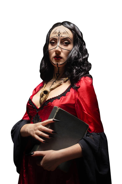 Odizolowana tajemnicza czarownica lub czarownica z runicznym makijażem w czerwonej sukience z drewnianym zwierzęcym amuletem czaszki trzymającym magiczną książkę. Koncepcja Halloween. - Zdjęcie, obraz