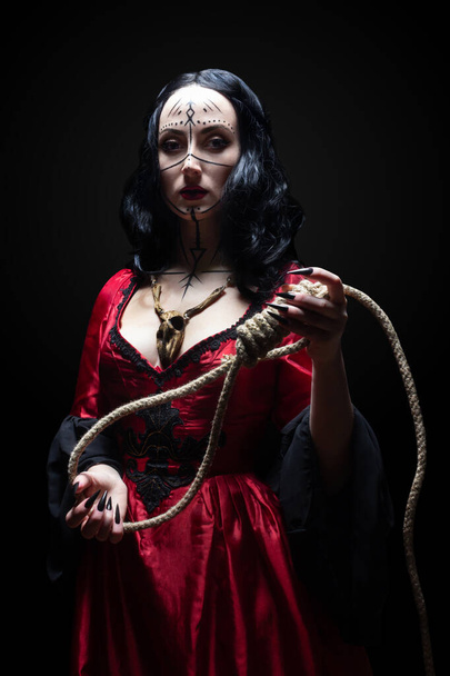 Beautiful brunette sorceress or witch with runic makeup in red gothic dress and wooden animal skull amulet holding a noose on black background. Halloween concept. - Photo, Image
