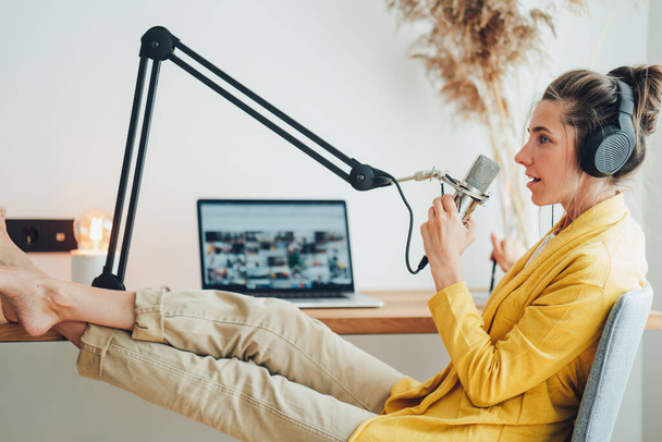 Woman sits with put your feet on table and recording podcast at his small cozy radio studio. Female podcaster streaming her voice into microphone - Fotoğraf, Görsel