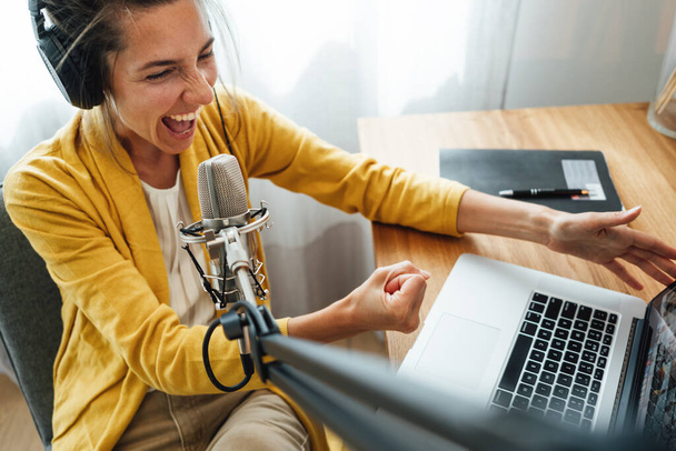 Charismatic woman radio host recording podcast at home studio. Female podcaster recording broadcasting into microphone using laptop - Foto, Bild