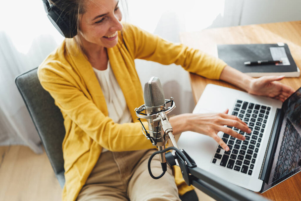Woman sits with put your feet on table and recording podcast at his small cozy radio studio. Female podcaster streaming her voice into microphone - 写真・画像