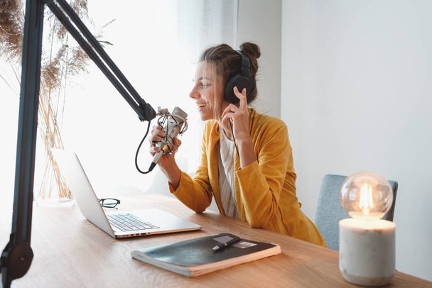 Young female radio host recording and broadcasting her podcast from homemade studio. Woman podcaster talk into microphone on table - Фото, изображение
